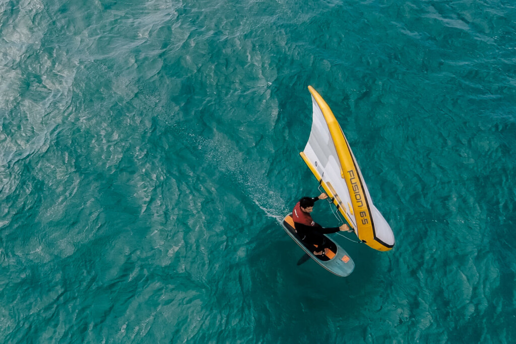Rideur de wingfoil vu du ciel sur une eau turquoise, avec une aile Ozone jaune et blanche